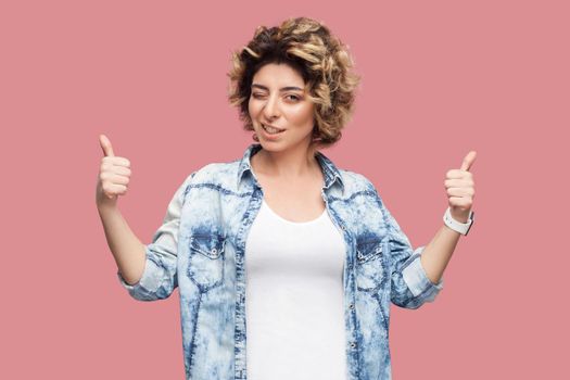 Portrait of happy satisfied young woman with curly hairstyle in casual blue shirt standing, thumbs up, winking and looking at camera with toothy smile. indoor studio shot, isolated on pink background.