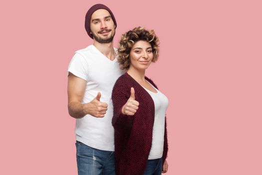 Portrait of satisfied couple of friends in casual style standing, hugging and showing thumbs up sign, looking at camera. Isolated, indoor, studio shot, pink background