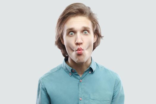 Portrait of funny handsome long haired blonde young man in blue casual shirt standing, looking at camera with big eyes and making fish lips. indoor studio shot, isolated on light grey background.