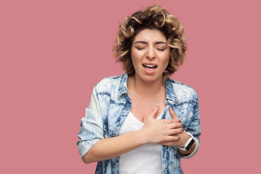 Heart attack or pain. Portrait of sick young woman with curly hairstyle in casual blue shirt standing and holding her painful herat and feeling bad. indoor studio shot, isolated on pink background.