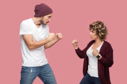 Side view portrait of upset angry couple of friends in casual style standing, disassembly and showing fists to each other, have conflict. Isolated, indoor, studio shot, pink background