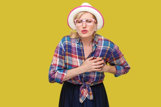 Heart attack pain or infarct. Portrait of mature woman in casual style with hat, eyeglasses standing, holding her breast because feeling pain on heart. indoor studio shot isolated on yellow background