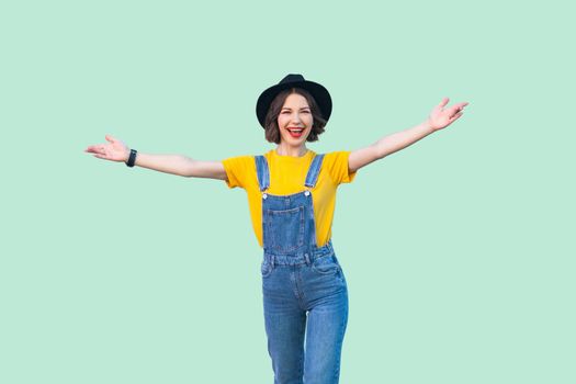 Portrait of happy pretty young girl in blue denim overalls, yellow shirt, black hat standing with raised arms and looking at camera with toothy smile. studio shot isolated on light green background.