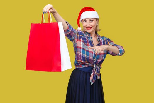 Happy modern middle aged woman in red santa cap and checkered shirt standing, holding and pointing finger to shopping bags and toothy smile, looking at camera. Indoor, studio shot, yellow background
