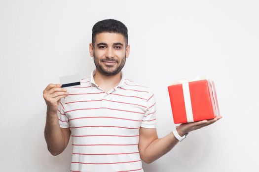 Satisfied attractive young adult man in t-shirt standing, holding big gift box and credit card with smile, online shopping, looking at camera. Studio shot, white background, isolated, indoor