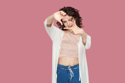 Portrait of beautiful brunette young woman with curly hairstyle in casual style standing and looking at camera with composition crop gesture hands. indoor studio shot isolated on pink background.