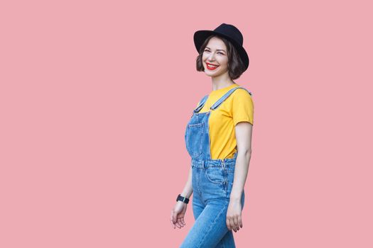 Portrait of happy beautiful young woman in yellow t-shirt and blue denim overalls with makeup and black hat standing and looking at camera with toothy smile. studio shot isolated on pink background.