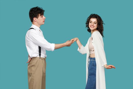 Profile side view portrait of brunette stylish gentleman standing and holding hand of cheerful happy young woman with toothy smile and happiness. indoor studio shot isolated on blue background.