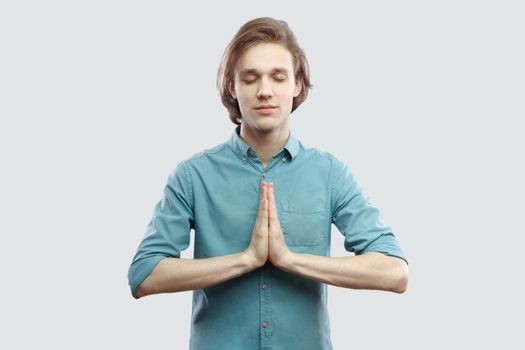 Portrait of calm handsome long haired blonde young man in blue casual shirt standing in yoga pose and meditating, and try to relax. indoor studio shot, isolated on light grey background.