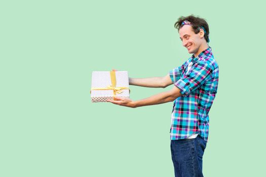 Side view of happy young hipster man in white t-shirt and checkered shirt standing, giving you present with yellow bow, toothy smile. Indoor, copy space, isolated, studio shot, green background