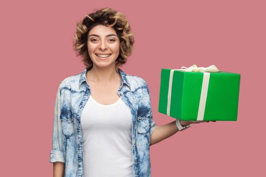 Portrait of happy beautiful young woman in blue shirt with curlty hairstyle standing and holding green gift box with toothy smile, looking at camera. Studio shot, pink background, isolated, indoor