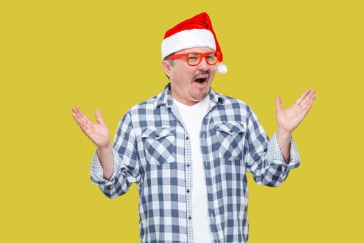 Portrait of funny modern middle aged man in red cap, eyeglasses and checkered shirt standing with hands up and surprised face looking at camera. Indoor, studio shot, isolated on yellow background