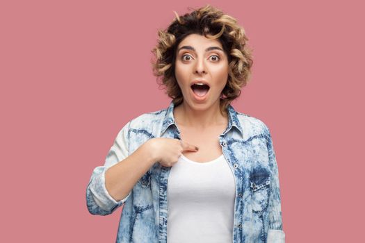 Portrait of shocked young woman with curly hairstyle in casual blue shirt standing, pointing herself and looking at camera with surprised face. indoor studio shot, isolated on pink background