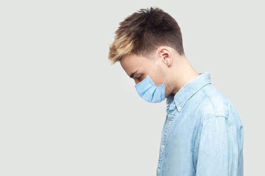 Profile side view portrait of sad upset alone young man with surgical medical mask in blue shirt standing, holding head down and depressed. indoor studio shot on grey background copy space.