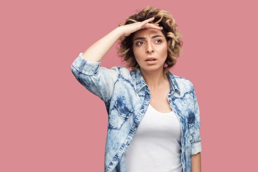 Portrait of attentive young woman with curly hairstyle in casual blue shirt standing with hand on forehead and looking away with serious face and attention. studio shot, isolated on pink background.