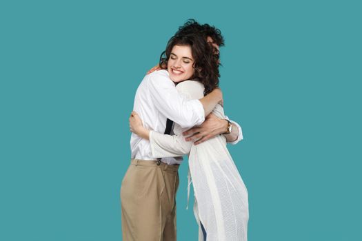 Profile side view portrait of two happy brunette stylish partner standing and hugging each other. love, friendship or partner greeting or celebration. indoor studio shot isolated on blue background.