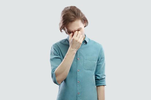 Portrait of sad alone handsome long haired blonde young man in blue casual shirt standing, holding his head down and crying. indoor studio shot, isolated on light grey background.