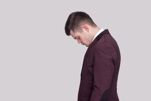 Profile side view portrait of sad handsome young man in violet suit and white shirt, standing and looking down with head down and bad mood. indoor studio shot, isolated on grey background.