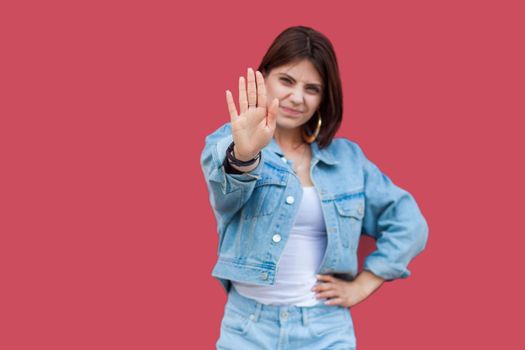Portrait of warning beautiful brunette young woman with makeup in denim casual style standing with hand on waist and looking at camera with stop sign hands. studio shot, isolated on red background.
