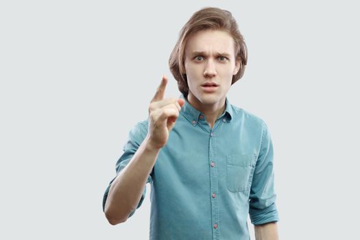 Portrait of serious handsome long haired blonde young man in blue casual shirt standing with warning sign and looking at camera. indoor studio shot, isolated on light grey background.