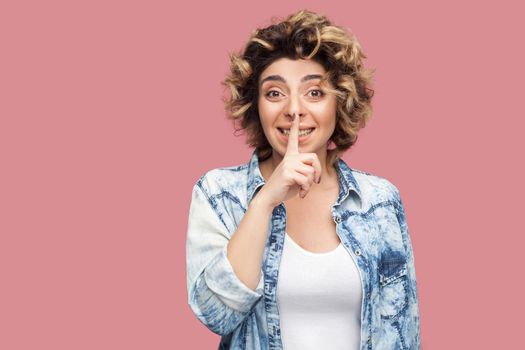 Portrait of worry young woman with curly hairstyle in casual blue shirt standing, telling secret and looking at camera with finger on lips secret sign. indoor studio shot, isolated on pink background.