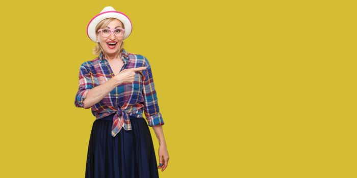 Portrait of surprised modern stylish mature woman in casual style with hat and eyeglasses standing, amazed looking at camera and pointing at wall. indoor studio shot isolated on yellow background.