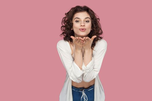 Portrait of happy in love beautiful brunette young woman with curly hairstyle in casual style standing and sending air kiss and looking at camera. indoor studio shot isolated on pink background.