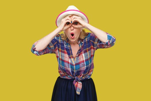 Portrait of funny surprised modern stylish mature woman in casual style with hat standing with binoculars gesture and looking with amazed face. indoor studio shot isolated on yellow background.