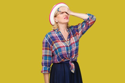 headache, mistake or problem. Portrait of sad disappointed stylish mature woman in casual style with hat standing with hand on forehead and upset. indoor studio shot isolated on yellow background.
