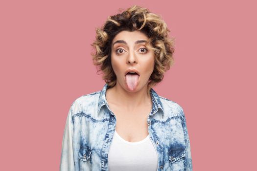 Portrait of crazy or shocked funny young woman with curly hairstyle in casual blue shirt standing, tongue out and looking at camera with big eyes. indoor studio shot, isolated on pink background.