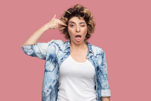Portrait of crazy young woman with curly hairstyle in casual blue shirt standing with pistol hand gun on her head and looking at camera. indoor studio shot, isolated on pink background.