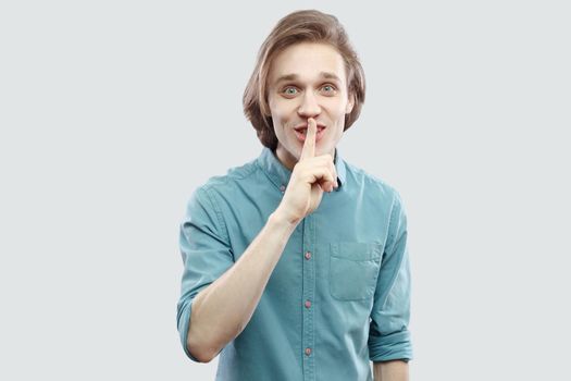 This is secret betwen us. Portrait of funny handsome long haired blonde young man in blue shirt standing looking at camera with silence sign. indoor studio shot, isolated on light grey background.
