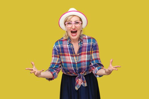 what do you want from me? Portrait of angry modern stylish mature woman in casual style with hat and eyeglasses standing, looking at camera and asking. indoor studio shot isolated on yellow background