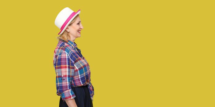 Profile side view portrait of happy successful modern stylish mature woman in casual style with hat standing, looking forward with toothy smile. indoor studio shot isolated on yellow background.