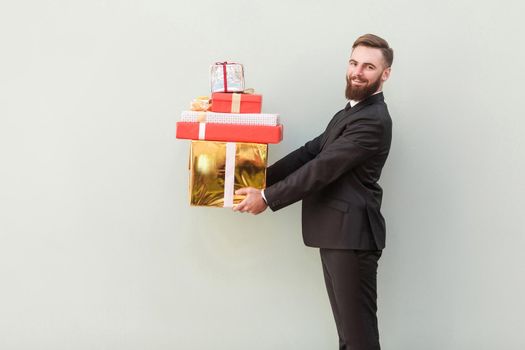 Profile, side view businessman toothy smile and holding gift boxes. Indoor shot, gray background