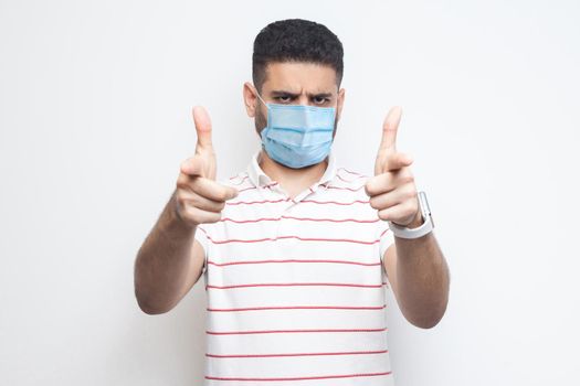Hey you. Portrait of serious bossy young man with surgical medical mask in striped t-shirt standing, looking and pointing at camera. indoor studio shot, isolated on white background.