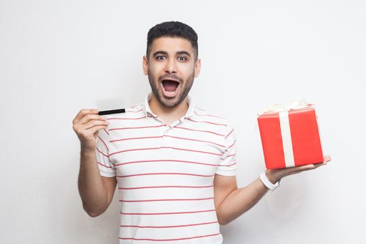 Unbelievable attractive young adult man in t-shirt standing, holding big gift box and credit card with shocked face, online shopping, looking at camera. Studio shot, white background, isolated, indoor