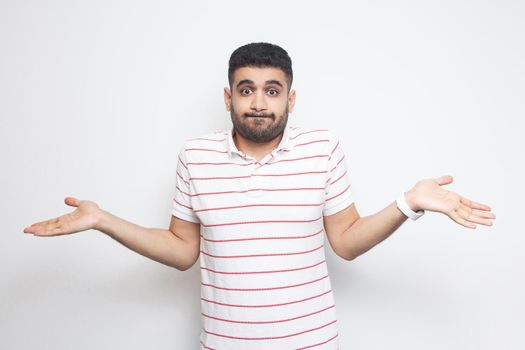 I don't know. Portrait of confused handsome bearded young man in striped t-shirt standing with raised arms and dont know what to do. indoor studio shot, isolated on white background.