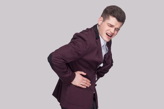 Profile side view portrait of unhappy handsome young man in violet suit and white shirt, standing and holding his painful stomach and feeling pain. indoor studio shot, isolated on grey background.