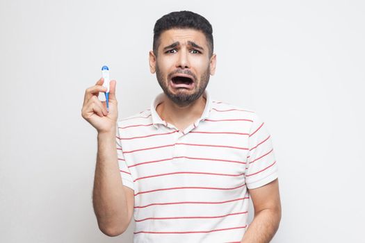 Scared worry sick young adult man in white t-shirt standing and holding thermometer are shoched for high temperature, looking at camera. Indoor, isolated, white background, studio shot