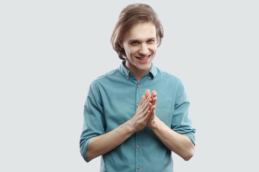 Portrait of cunning handsome long haired blonde young man in blue casual shirt standing and looking at camera with bitchy plan. indoor studio shot, isolated on light grey background.