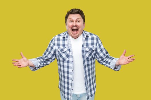 Portrait of surprised handsome middle aged businessman in casual checkered shirt standing with raised arms, looking at camera with unbelievable amazed face. studio shot, isolated on yellow background.