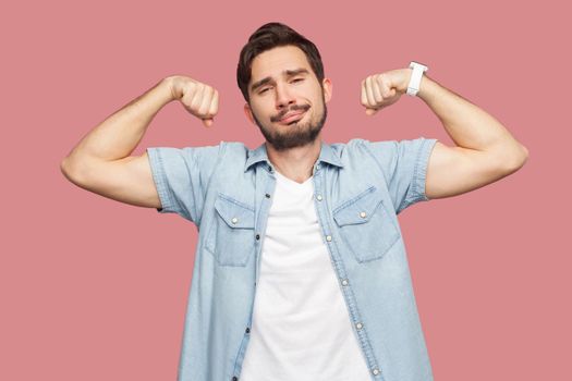 Portrait of proud satisfied handsome bearded young man in blue casual style shirt standing in body building pose and showing his strongness. indoor studio shot, isolated on pink background.
