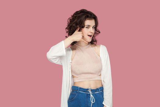Portrait of funny beautiful brunette young woman with curly hairstyle in casual style standing with call hands gesture and looking at camera. indoor studio shot isolated on pink background.