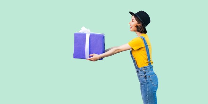Side view of happy beautiful young girl in hipster wear in denim overalls and black hat standing and giving you big heavy gift box with toothy smile. Studio shot, green background, isolated