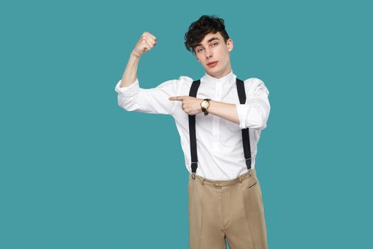 Proud man pointing at his biceps and looking at camera. Portrait of handsome hipster curly young businessman in classic casual white shirt and suspender. indoor studio shot isolated on blue background