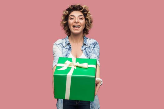 Portrait of happy beautiful young woman in blue shirt with curlty hairstyle standing and giving you green gift box with toothy smile, looking at camera. Studio shot, pink background, isolated, indoor