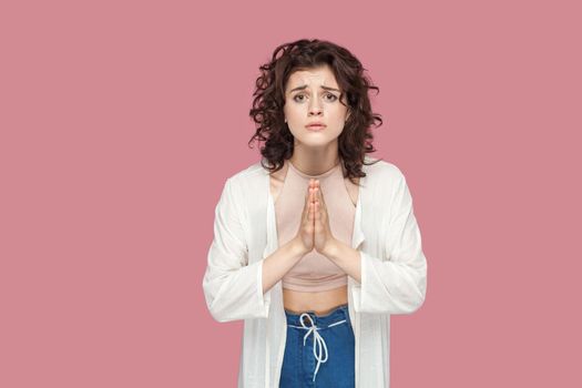 Please help me or give me one chance more. Portrait of hopeful young woman with curly hairstyle in casual style standing with palm hands and looking at camera. studio shot isolated on pink background.