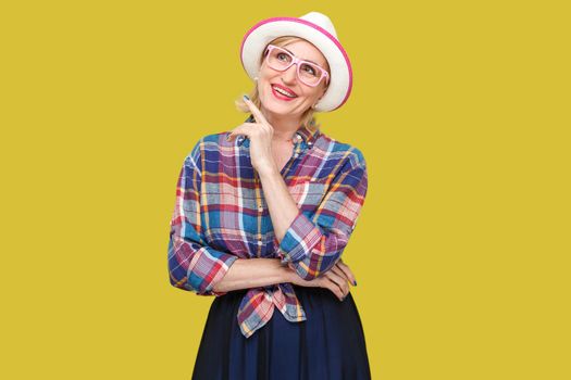 dreaming about something beautiful. Portrait of toothy smiley modern stylish mature woman in casual style with hat standing, looking away and dreaming. indoor studio shot isolated on yellow background