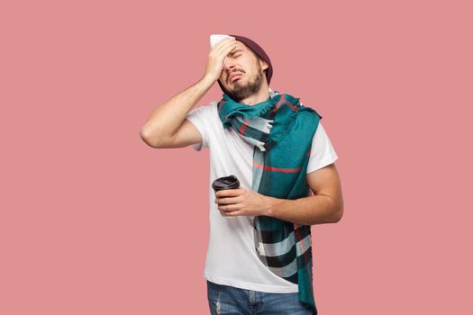 Sickness modern bearded young hipster man in white shirt, hat and scarf standing, touching with hand his head, have strong headache, holding hot tea in cup. Indoor,isolated,studio shot,pink background